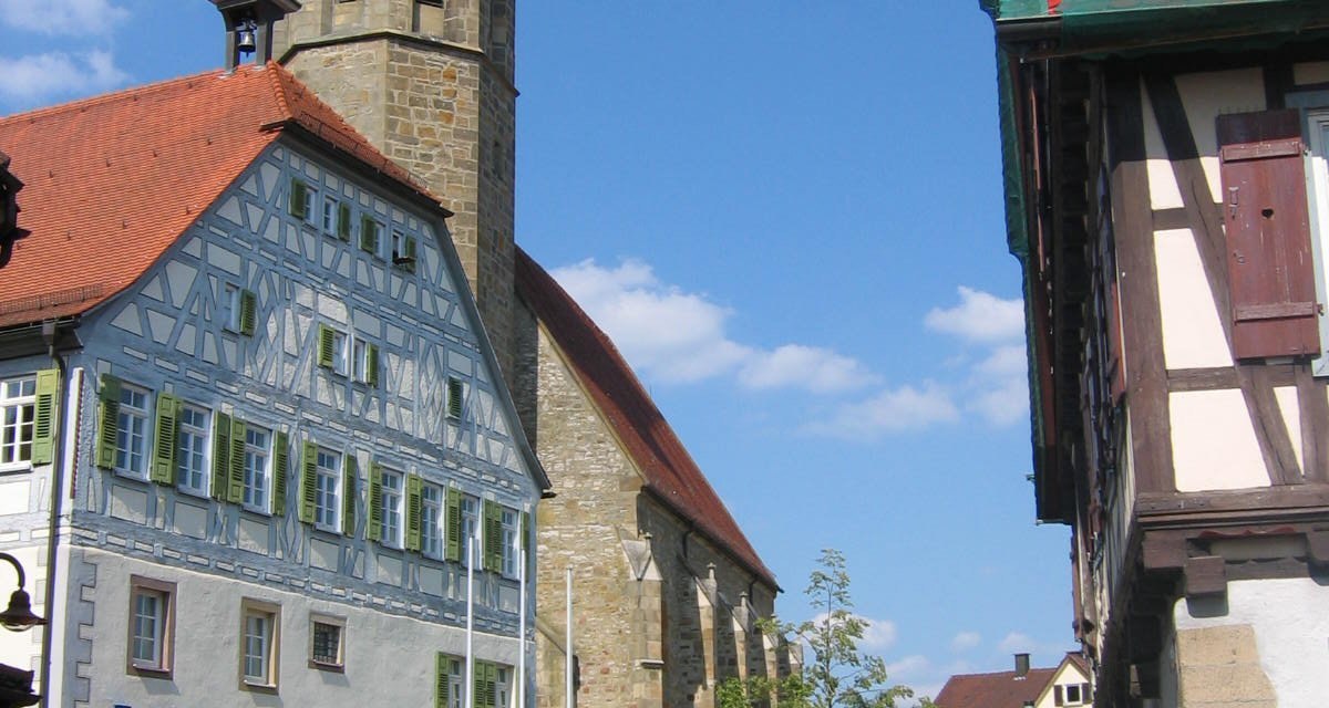 Fachwerkgeb&auml;ude mit Kirchturm und Uhr, daneben ein rotes Auto. Blauer Himmel und gepflasterte Stra&szlig;e im Vordergrund., &copy; Land der 1000 H&uuml;gel - Kraichgau-Stromberg