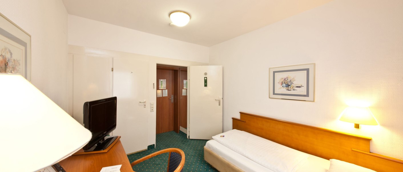Ein einfach eingerichtetes Einzelzimmer mit Einzelbett, Schreibtisch, Fernseher und einem Bild an der Wand. Der Boden ist mit grünem Teppich ausgelegt., © Novum Boulevard Stuttgart City