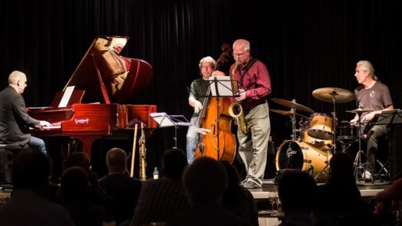 Eine Jazzband spielt auf einer B&uuml;hne. Zu sehen sind ein Pianist, ein Kontrabassist, ein Saxophonist und ein Schlagzeuger. Das Publikum sitzt im Vordergrund., &copy; Carl Clements & RJT [Real Jazz Trio]