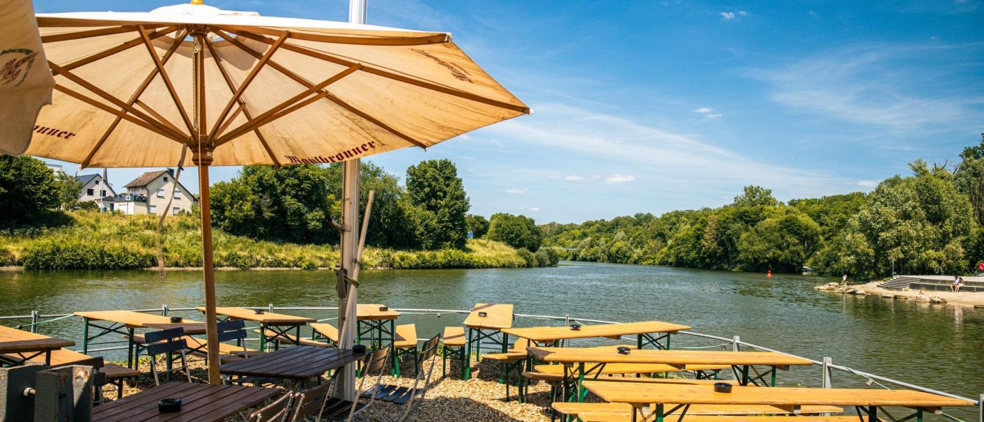 Ein Biergarten mit Holztischen und -stühlen unter einem großen Sonnenschirm am Ufer eines Flusses, umgeben von grüner Landschaft., © Stuttgart-Marketing GmbH, Sarah Schmid