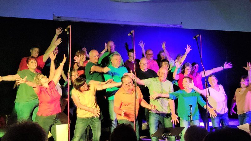 Ein Chor in bunten Outfits singt und tanzt auf einer B&uuml;hne mit lebendiger Beleuchtung.