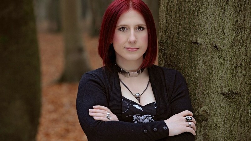 Eine Person mit roten Haaren steht mit verschr&auml;nkten Armen vor einem Baum in einem herbstlichen Wald., &copy; Scala Kultur Live gGmbH