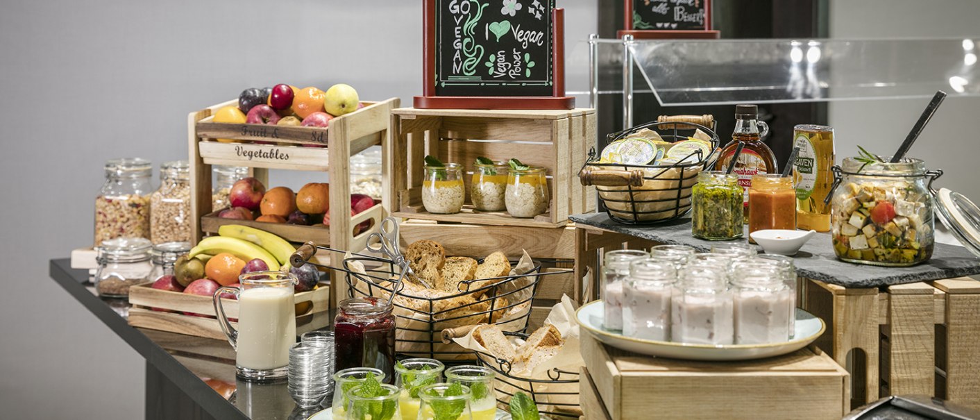 Ein Frühstücksbuffet mit Obst, Joghurt, Brot, Aufstrichen und Getränken. Ein Schild weist auf eine vegane Ecke hin., © Arcotel Hotels