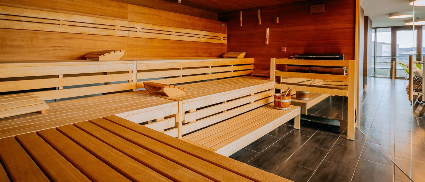 Moderne Sauna mit Holzbänken, einem Eimer und einem Fenster mit Blick nach draußen. Entspannte Atmosphäre im Spa-Bereich., © V8 HOTEL Moderne Sauna mit Holzbänken, einem Eimer und einem Fenster mit Blick nach draußen. Entspannte Atmosphäre im Spa-Bereich., © V8 HOTEL
