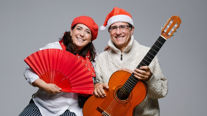 Ein Mann und eine Frau tragen Weihnachtsmützen. Die Frau hält einen roten Fächer, der Mann eine Gitarre. Beide lächeln in die Kamera., © Duo Alegria Ein Mann und eine Frau tragen Weihnachtsmützen. Die Frau hält einen roten Fächer, der Mann eine Gitarre. Beide lächeln in die Kamera., © Duo Alegria