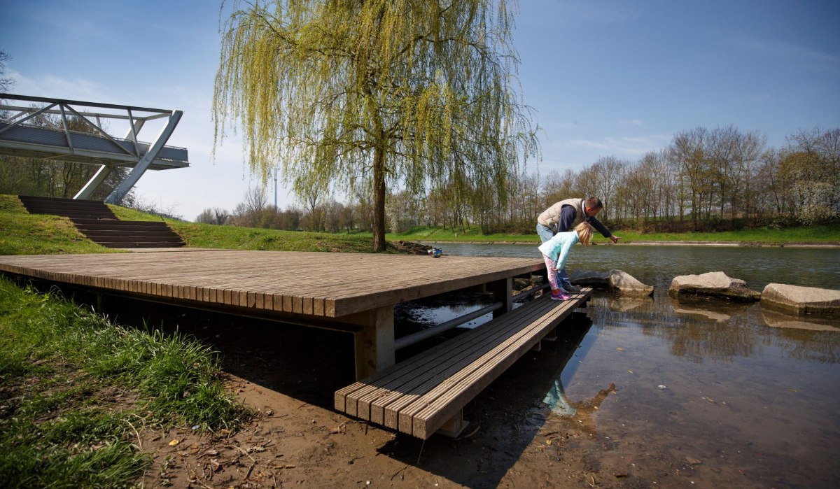 Ein Mann und ein Kind auf einem Holzsteg am Flussufer, umgeben von Natur. Eine moderne Brücke und ein Baum im Hintergrund., © Stoppel Ein Mann und ein Kind auf einem Holzsteg am Flussufer, umgeben von Natur. Eine moderne Brücke und ein Baum im Hintergrund., © Stoppel