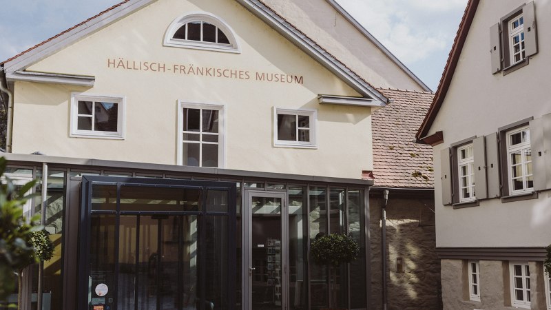 Eingang des Hällisch-Fränkischen Museums in Schwäbisch Hall, mit moderner Glasfassade und historischen Gebäuden im Hintergrund., © Nico Kurth