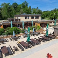 Au&szlig;enbereich von Weitmanns Waldhaus mit Holztischen, B&auml;nken und Sonnenschirmen, umgeben von gr&uuml;nen B&auml;umen und blauen Himmel., &copy; Timo Weitmann