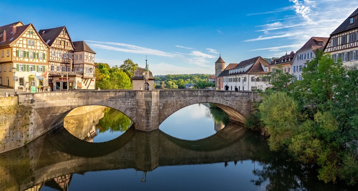 Die Henkersbr&uuml;cke in Schw&auml;bisch Hall, umgeben von Fachwerkh&auml;usern, spiegelt sich im ruhigen Wasser unter einem klaren blauen Himmel., &copy; Michael K&uuml;hneisen