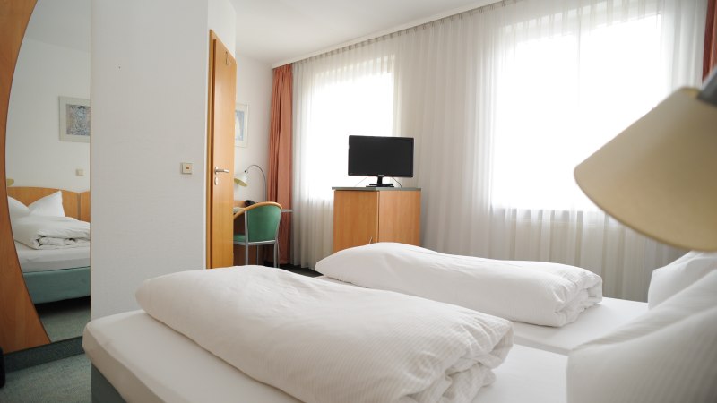 Ein Hotelzimmer mit zwei Einzelbetten, einem Fernseher auf einem Schrank, einem Schreibtisch mit Stuhl und großen Fenstern mit Vorhängen., © TOMAS