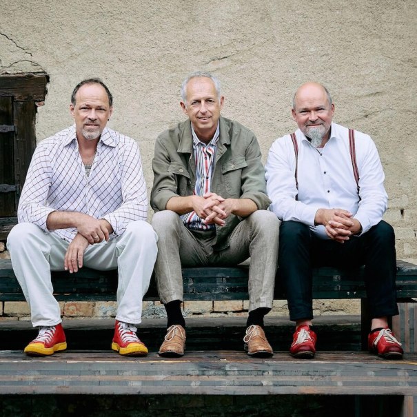 Drei Männer sitzen auf einer Bank vor einer rustikalen Wand. Sie tragen bunte Schuhe und lässige Kleidung, die eine entspannte Atmosphäre vermittelt., © Mauerwerk GmbH