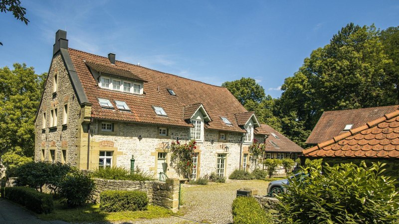 Ein charmantes, historisches Gebäude mit rotem Ziegeldach und blühenden Rosen an der Fassade, umgeben von üppigem Grün und einem gepflasterten Weg., © SMG, Sarah Schmid Ein charmantes, historisches Gebäude mit rotem Ziegeldach und blühenden Rosen an der Fassade, umgeben von üppigem Grün und einem gepflasterten Weg., © SMG, Sarah Schmid