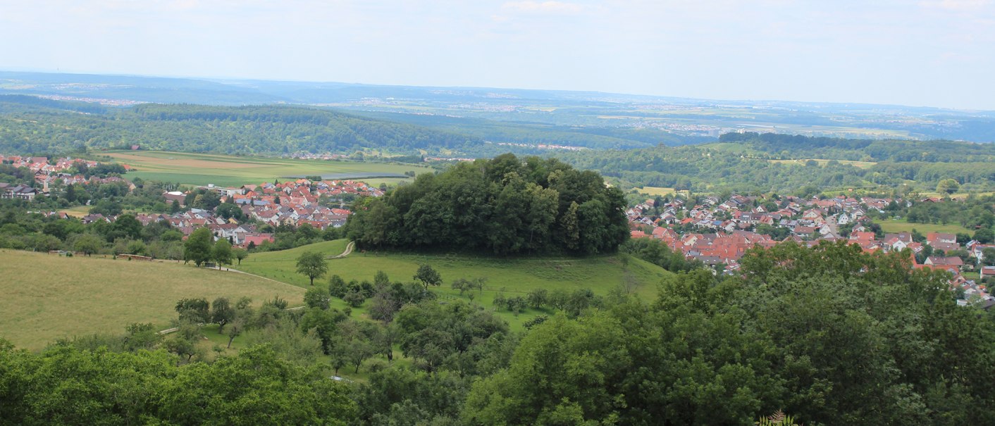 Blick auf Beuren mit einem bewaldeten Hügel im Vordergrund und einem Dorf mit roten Dächern im Hintergrund, umgeben von grüner Landschaft., © Kurverwaltung Beuren Blick auf Beuren mit einem bewaldeten Hügel im Vordergrund und einem Dorf mit roten Dächern im Hintergrund, umgeben von grüner Landschaft., © Kurverwaltung Beuren