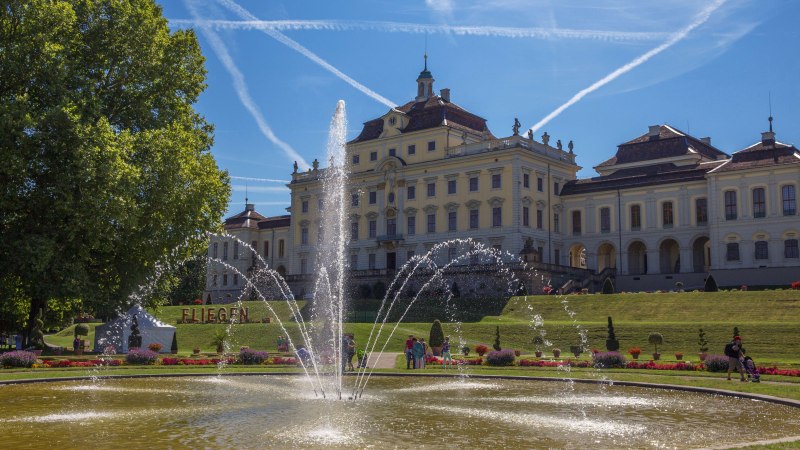 Schloss Ludwigsburg mit prächtigem Garten und Springbrunnen, umgeben von blühenden Blumen. Der Himmel ist blau mit Kondensstreifen., © Tourismus & Events Ludwigsburg
