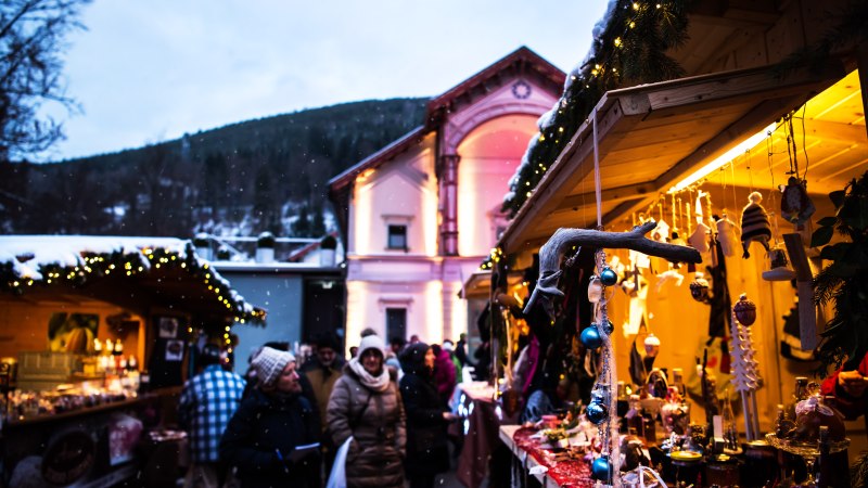 Weihnachtsmarkt mit beleuchteten Ständen, Dekorationen und Menschen in Winterkleidung. Im Hintergrund ein schneebedeckter Hügel und ein beleuchtetes Gebäude., © Locher Fotodesign&Manufaktur