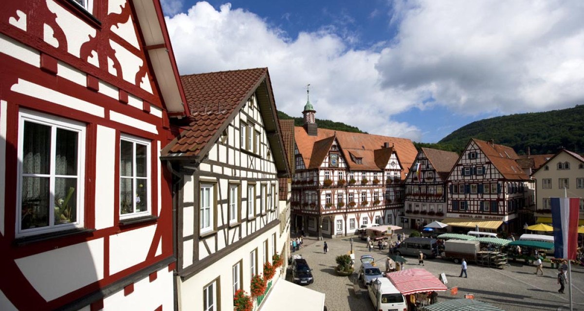 Blick auf den Marktplatz in Bad Urach mit malerischen Fachwerkhäusern und bunten Marktständen unter einem blauen Himmel mit Wolken., © Bad Urach Tourismus Blick auf den Marktplatz in Bad Urach mit malerischen Fachwerkhäusern und bunten Marktständen unter einem blauen Himmel mit Wolken., © Bad Urach Tourismus