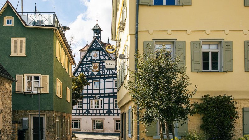 Blick auf ein historische Fachwerkhaus in Bad Überkingen, eingerahmt von bunten Gebäuden, dem Rathaus und einem Baum im Vordergrund., © SMG, Sarah Schmid Blick auf ein historische Fachwerkhaus in Bad Überkingen, eingerahmt von bunten Gebäuden, dem Rathaus und einem Baum im Vordergrund., © SMG, Sarah Schmid