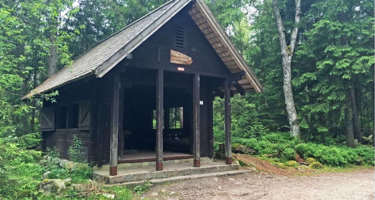 Eine kleine Holzh&uuml;tte im Wald, umgeben von hohen B&auml;umen. &Uuml;ber dem Eingang h&auml;ngt ein Schild. Der Boden ist mit Kies bedeckt., &copy; www.pro-cycl.de