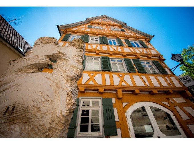 Fachwerkhaus in Schorndorf mit gelben und weißen Balken, grünen Fensterläden und einer Holzskulptur im Vordergrund., © Eigenbetrieb Tourismus und Citymanagement Schorndorf Fachwerkhaus in Schorndorf mit gelben und weißen Balken, grünen Fensterläden und einer Holzskulptur im Vordergrund., © Eigenbetrieb Tourismus und Citymanagement Schorndorf