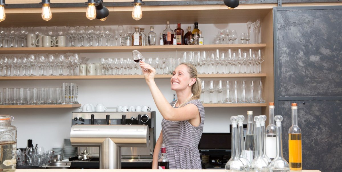 Frau in einem Caf&eacute; pr&uuml;ft ein Weinglas. Im Hintergrund sind viele Gl&auml;ser und Flaschen auf Regalen. Eine Kaffeemaschine steht auf der Theke.