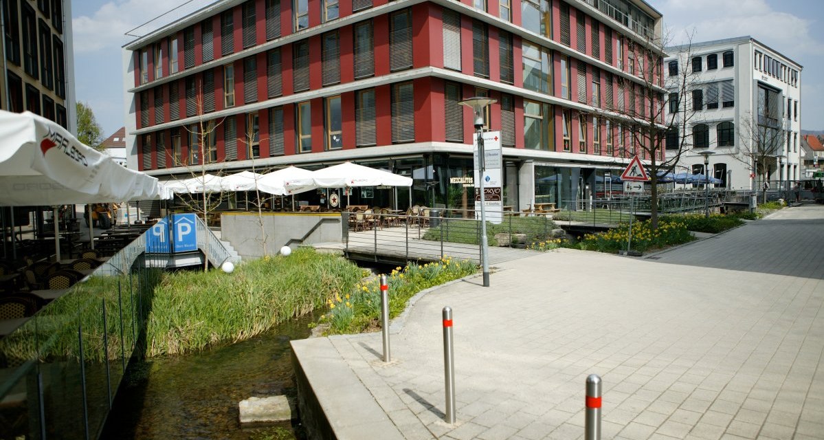 Modernes Gebäude mit roten Fassaden, Glasfronten und Außenterrasse. Ein kleiner Bach fließt daneben, umgeben von Gehweg und Blumen., © Sphäre Verlag Modernes Gebäude mit roten Fassaden, Glasfronten und Außenterrasse. Ein kleiner Bach fließt daneben, umgeben von Gehweg und Blumen., © Sphäre Verlag
