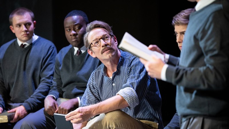 Schauspieler in Schuluniformen und ein Mann in Freizeitkleidung sitzen auf einer B&uuml;hne und lesen aus B&uuml;chern.