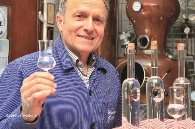 Mann in blauer Jacke h&auml;lt ein Glas mit klarer Fl&uuml;ssigkeit vor einer Destillationsanlage. Auf dem Tisch stehen Flaschen., &copy; Eigenbetrieb Tourismus und Citymanagement Schorndorf