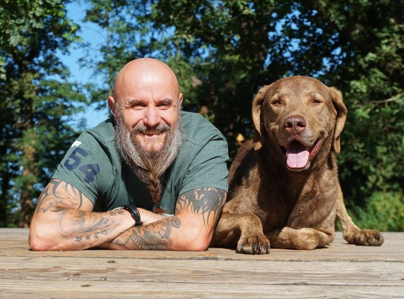 Ein Mann mit Bart und Tattoos liegt neben einem braunen Hund auf einem Holzdeck. Beide lächeln in die Kamera, umgeben von grünen Bäumen., © Stadthalle Leonberg Ein Mann mit Bart und Tattoos liegt neben einem braunen Hund auf einem Holzdeck. Beide lächeln in die Kamera, umgeben von grünen Bäumen., © Stadthalle Leonberg