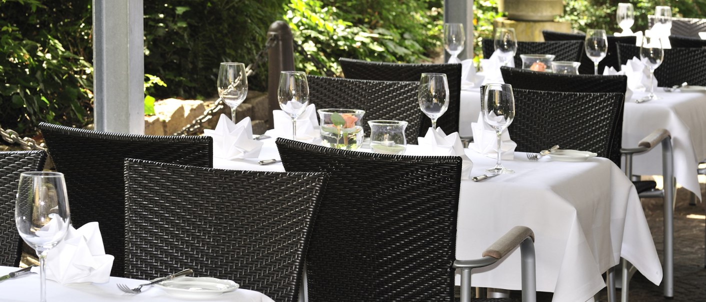 Elegante Restaurantterrasse mit gedeckten Tischen, wei&szlig;en Tischdecken und Weingl&auml;sern, umgeben von gr&uuml;ner Vegetation., &copy; cSchupfnudl