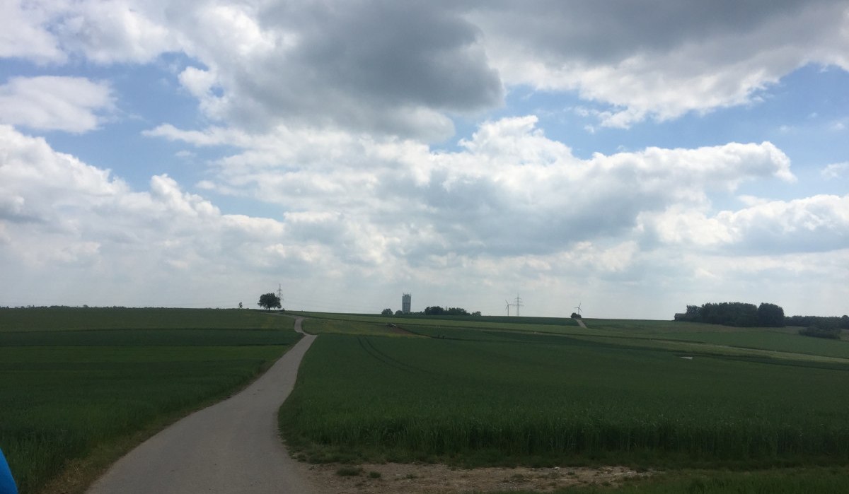 Ein Feldweg führt durch grüne Felder zu einem fernen Wasserturm und Windrädern, unter einem bewölkten Himmel., © www.pro-cycl.de Ein Feldweg führt durch grüne Felder zu einem fernen Wasserturm und Windrädern, unter einem bewölkten Himmel., © www.pro-cycl.de