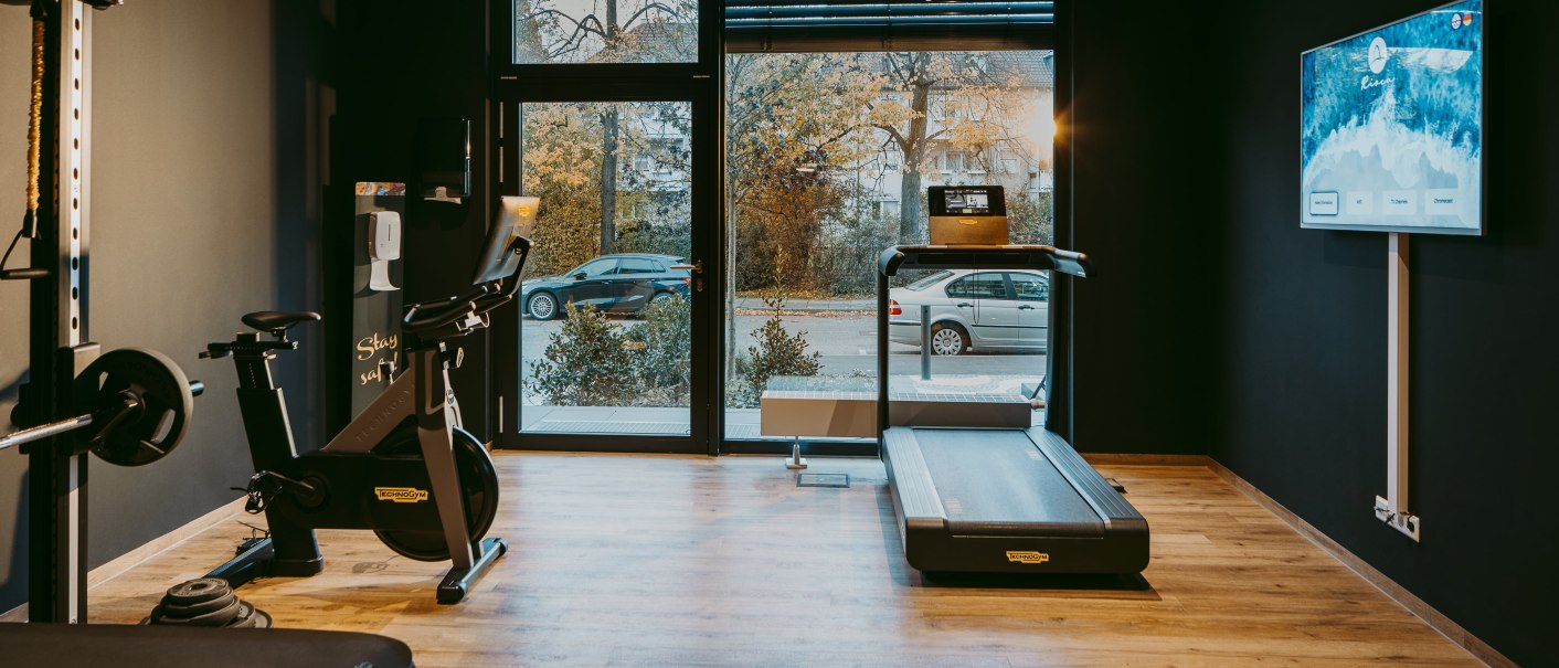 Moderner Fitnessraum mit Laufband, Fahrrad und Hanteln. Große Fenster bieten Blick auf die Straße. Ein Bildschirm hängt an der Wand., © Rioca Stuttgart Posto 4 GmbH