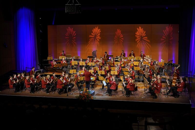 Ein Orchester in roten Uniformen spielt auf einer Bühne. Der Hintergrund ist mit bunten Lichteffekten dekoriert., © Feuerwehr-Musikkapelle Dagersheim e. V.