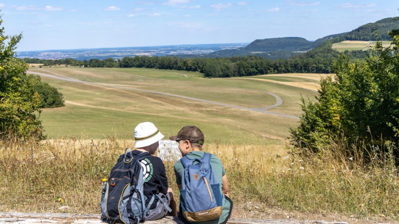 Zwei Personen mit Rucks&auml;cken sitzen auf einem Baumstamm und blicken auf die Landebahn des Segelflugplatzes Hornberg in einer gr&uuml;nen, h&uuml;geligen Landschaft., &copy; Foto Thomas Zehnder