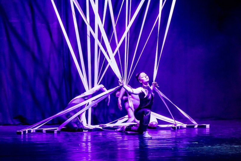 Zwei Tänzerinnen in violettem Licht, umgeben von weißen Bändern, auf einer Bühne. Eine kniet, die andere ist teils verdeckt. Dramatische Beleuchtung., © Stadthalle Leonberg