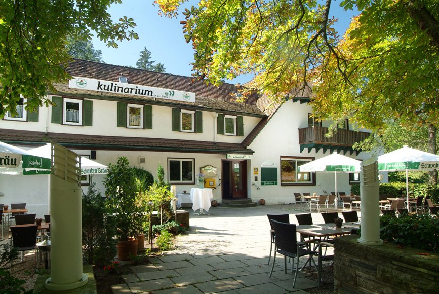 Ein Restaurant mit Terrasse, umgeben von Bäumen. Tische und Stühle stehen unter Sonnenschirmen. Das Gebäude hat grüne Fensterläden., © Kulinarium an der Glems