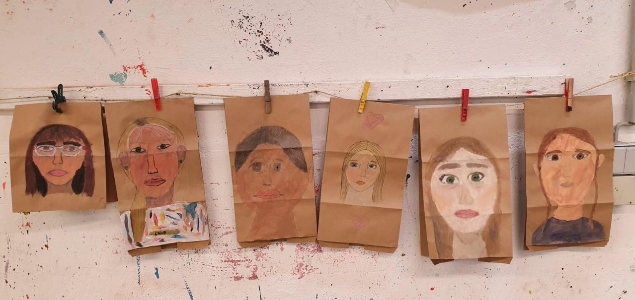 Sechs gemalte Selbstportraits von Kindern auf braunem Papier, aufgehängt mit bunten Wäscheklammern an einer weißen, bemalten Wand., © Kunstschule Labyrinth Sechs gemalte Selbstportraits von Kindern auf braunem Papier, aufgehängt mit bunten Wäscheklammern an einer weißen, bemalten Wand., © Kunstschule Labyrinth