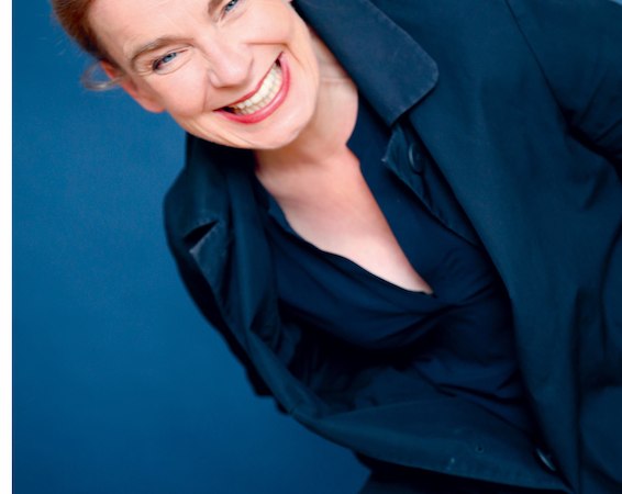 Eine Frau in einem schwarzen Mantel lacht herzlich vor einem blauen Hintergrund., © Dieselstrasse Kulturzentrum e.V.