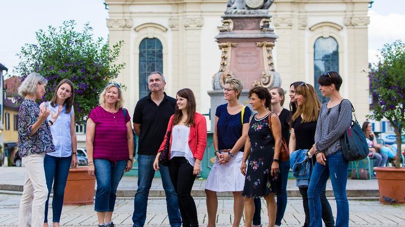 Eine Gruppe von Menschen steht vor einem historischen Gebäude in Ludwigsburg, während eine Frau spricht. Im Hintergrund ist eine Statue zu sehen., © © T&E, Benjamin Stollenberg Eine Gruppe von Menschen steht vor einem historischen Gebäude in Ludwigsburg, während eine Frau spricht. Im Hintergrund ist eine Statue zu sehen., © © T&E, Benjamin Stollenberg