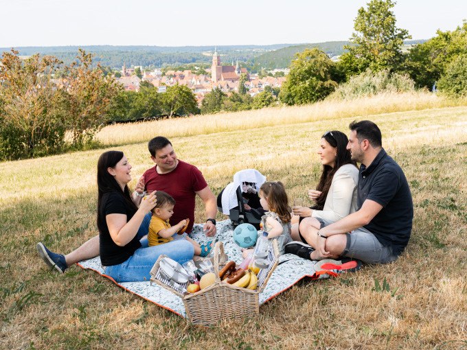 Familienpicknick auf einer Wiese mit Blick auf eine Stadt. Erwachsene und Kinder sitzen auf einer Decke mit einem Picknickkorb voller Snacks., &copy; Stadtverwaltung Weil der Stadt