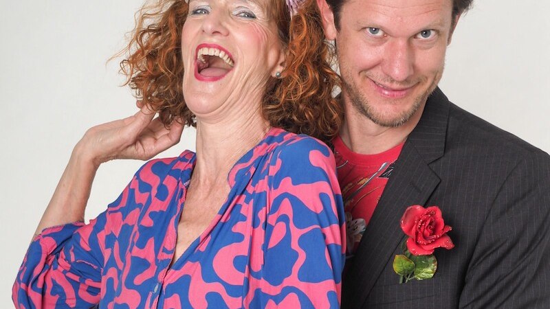 Eine Frau in einem pink-blauen Kleid lacht herzlich, neben ihr steht ein Mann im Anzug mit einer roten Blume im Knopfloch., © Mauerwerk GmbH Eine Frau in einem pink-blauen Kleid lacht herzlich, neben ihr steht ein Mann im Anzug mit einer roten Blume im Knopfloch., © Mauerwerk GmbH