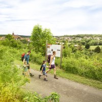 Eine Familie wandert auf einem Weg, vorbei an einer Informationstafel. Im Hintergrund ist eine Stadt mit Kirche und grüner Landschaft zu sehen., © Markus Born Eine Familie wandert auf einem Weg, vorbei an einer Informationstafel. Im Hintergrund ist eine Stadt mit Kirche und grüner Landschaft zu sehen., © Markus Born
