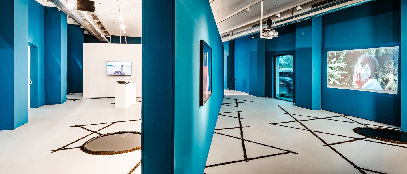 Moderne Galerie mit blauen Wänden, Videoinstallationen und geometrischen Mustern auf dem Boden. Industrielles Design mit sichtbaren Lüftungsrohren., © Ifa Galerie Stuttgart