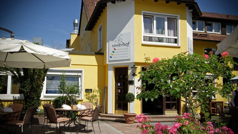Gelbes Gebäude mit Terrasse, Tischen und Stühlen. Sonnenschirme und blühende Pflanzen im Vordergrund. Schild mit der Aufschrift 'römerhof' an der Fassade., © Römerhof Hotelbetriebs GmbH