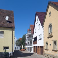 Eine ruhige Straße mit Häusern und einem klaren blauen Himmel. Die Gebäude haben rote Ziegeldächer und helle Fassaden., © Petra Natzkowski Eine ruhige Straße mit Häusern und einem klaren blauen Himmel. Die Gebäude haben rote Ziegeldächer und helle Fassaden., © Petra Natzkowski