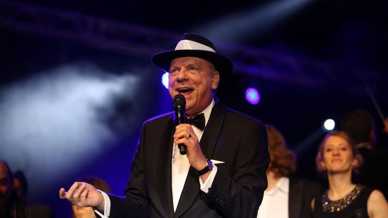 Ein Mann im Anzug und Hut singt auf einer Bühne. Er hält ein Mikrofon und wird von einem Orchester begleitet. Die Beleuchtung ist stimmungsvoll., © Touristik Bad Wildbad GmbH Ein Mann im Anzug und Hut singt auf einer Bühne. Er hält ein Mikrofon und wird von einem Orchester begleitet. Die Beleuchtung ist stimmungsvoll., © Touristik Bad Wildbad GmbH