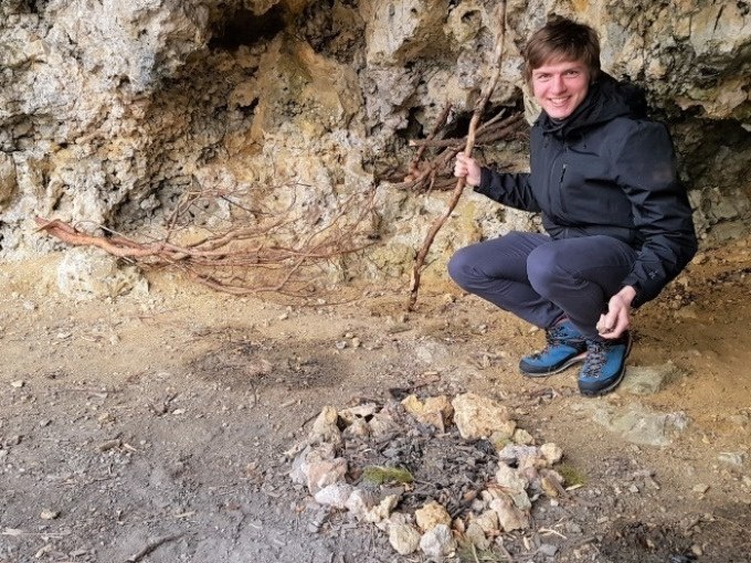 Eine Person in schwarzer Jacke kniet in einer Höhle vor einem Steinkreis. Sie hält einen Stock und lächelt in die Kamera., © SwabianTravel Eine Person in schwarzer Jacke kniet in einer Höhle vor einem Steinkreis. Sie hält einen Stock und lächelt in die Kamera., © SwabianTravel