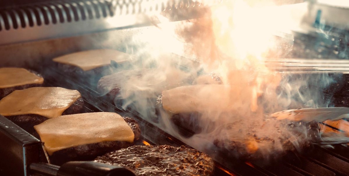 Burger-Patties mit geschmolzenem Käse auf einem Grill, umgeben von Rauch und Flammen., © Bady's