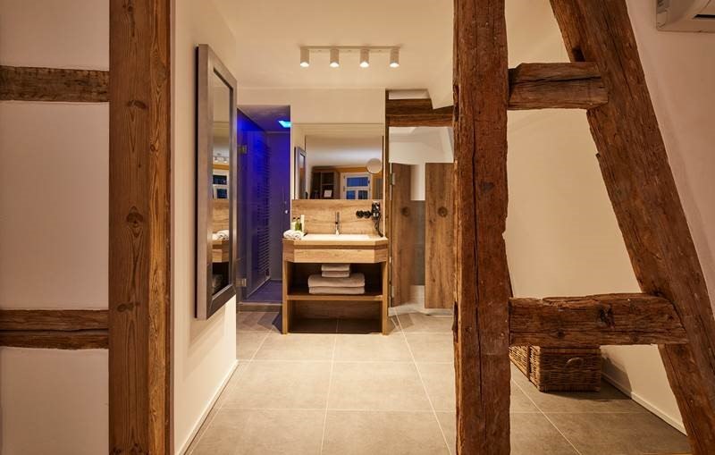 Modernes Badezimmer mit sichtbaren Holzbalken, einem Waschtisch mit Spiegel und Handtüchern. Beleuchtung und blauer Lichtakzent im Hintergrund., © Hotel Restaurant Bachofer Modernes Badezimmer mit sichtbaren Holzbalken, einem Waschtisch mit Spiegel und Handtüchern. Beleuchtung und blauer Lichtakzent im Hintergrund., © Hotel Restaurant Bachofer