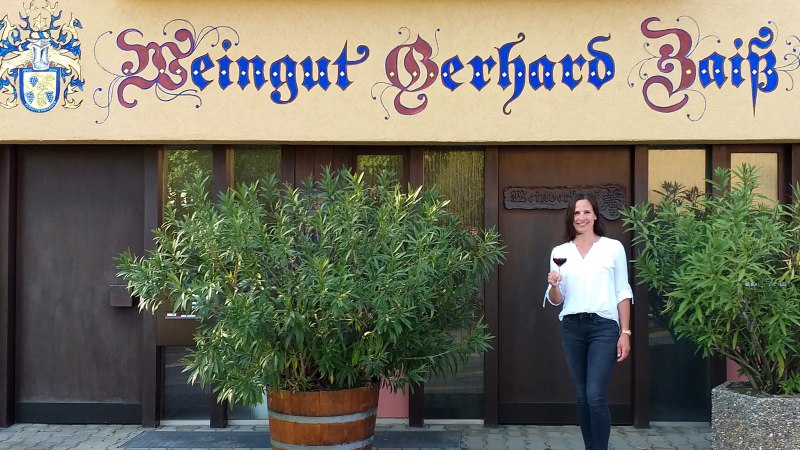 Eine Frau steht vor dem Weingut Gerhard Zaiß, hält ein Glas Rotwein und lächelt. Das Gebäude hat eine dekorative Beschriftung und Pflanzen im Vordergrund., © Simone Mack