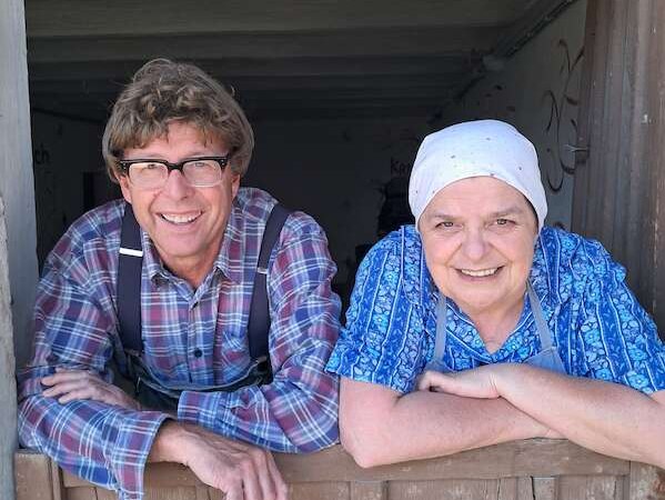 Zwei Personen lehnen sich lächelnd über eine Holztür. Eine Person trägt ein kariertes Hemd, die andere ein blaues Kopftuch und eine Schürze., © SBEntertainment GmbH & Co. KG